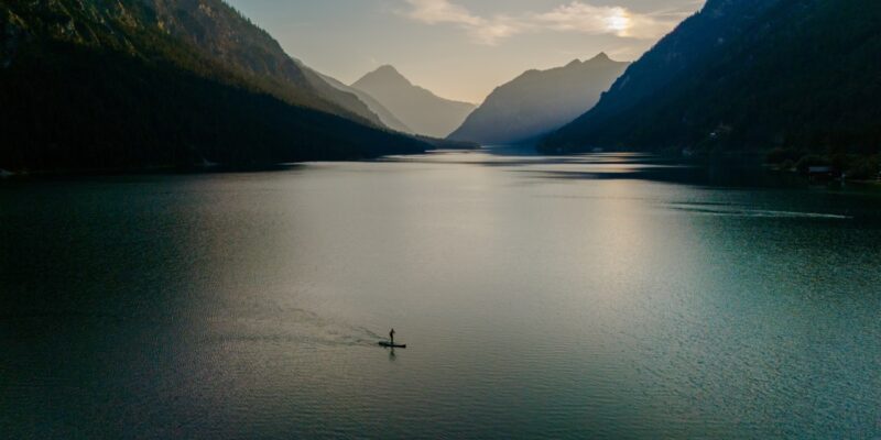 A lone paddleboarder glides over Plansee's calm waters at sunrise, as the sun casts a warm glow on the lake and highlights the stunning mountains surrounding this serene oasis.